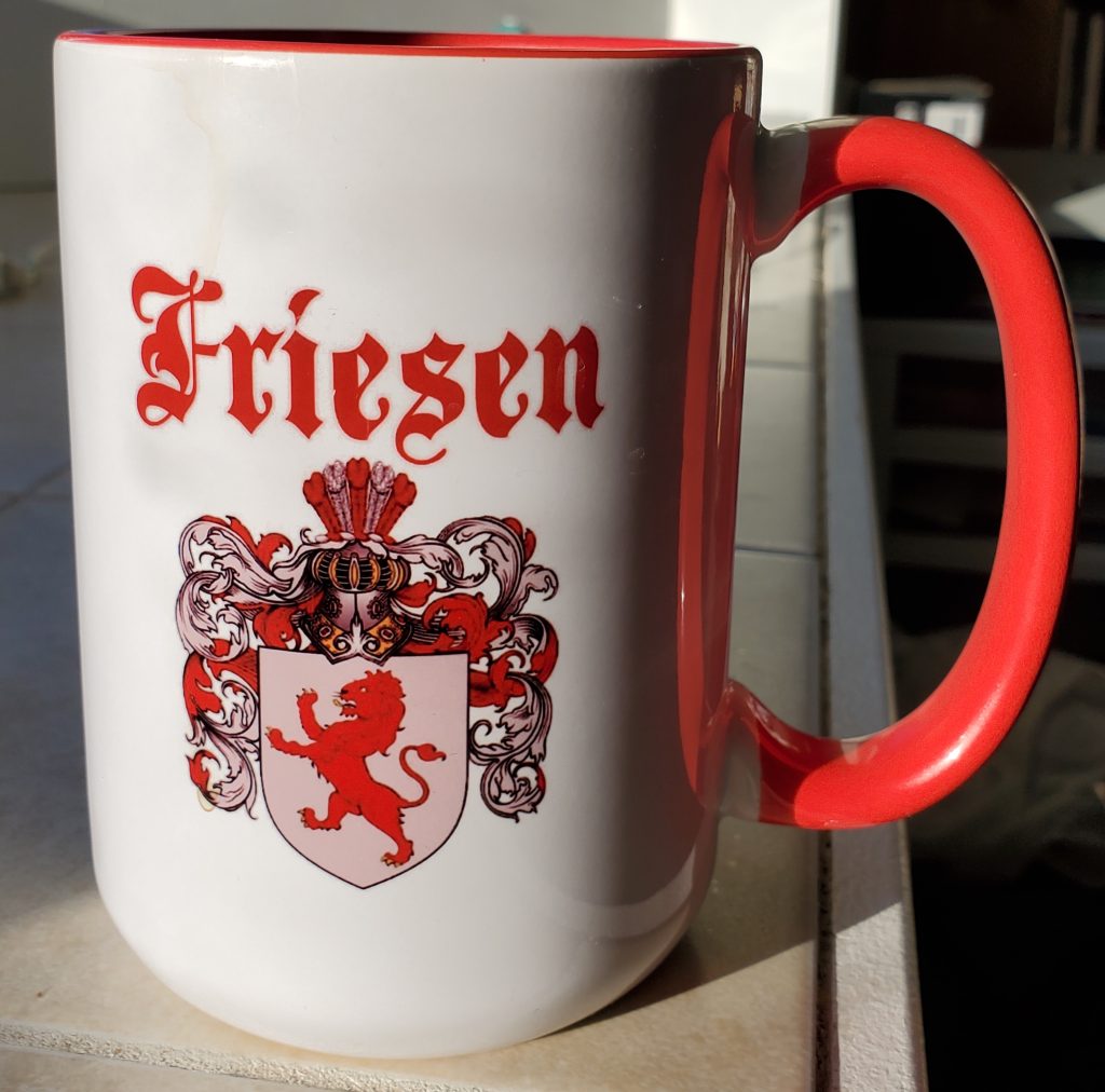 A white mug with a red handle. In fancy red script is written Friesen, with a coat of arms beneath.